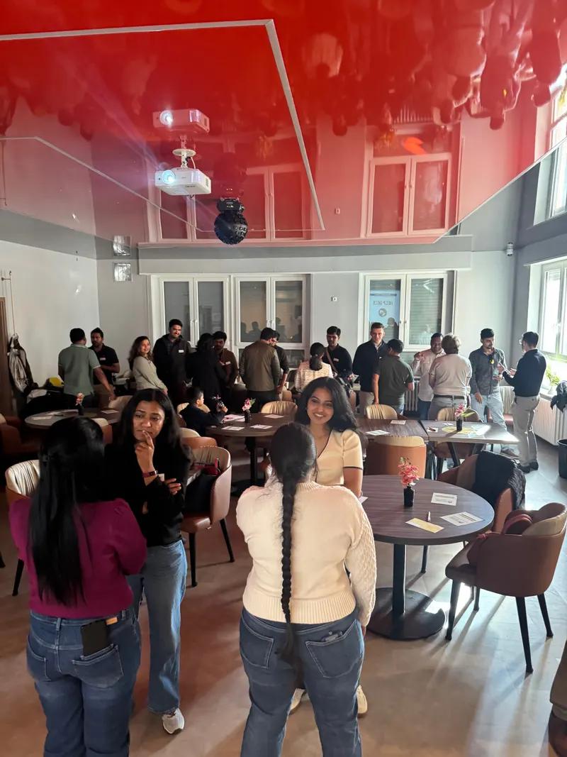 Attendees stand and chat in small groups around tables in a venue with a red ceiling.