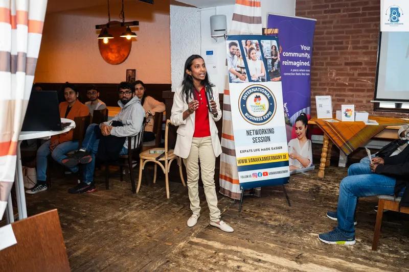 A woman in a blazer speaks while standing next to a networking session banner, with seated attendees nearby in a rustic-style venue.