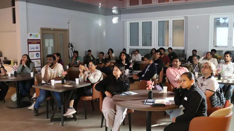 Rows of attendees sit at round tables listening during a seminar in a meeting room.