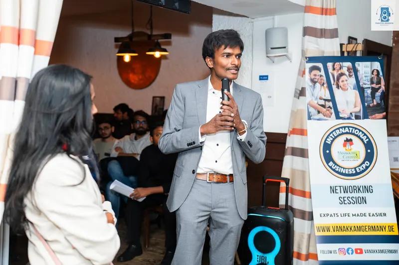 A man in a grey suit holds a microphone and speaks, standing next to a roll-up banner for an international business expo networking session.
