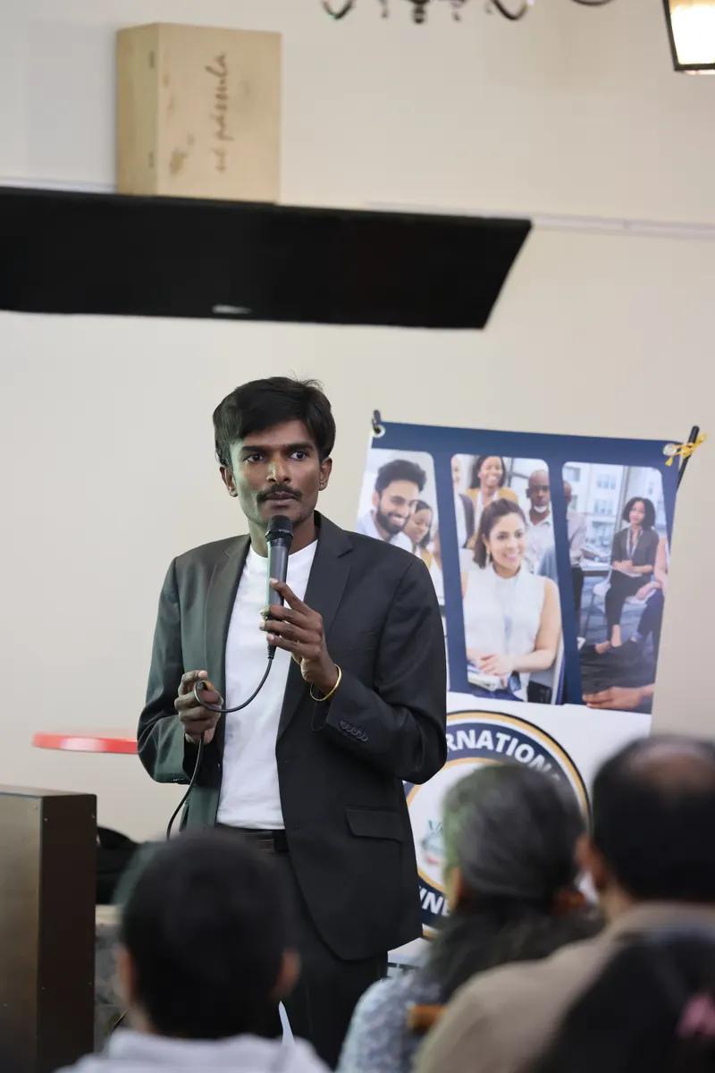 A man in a dark blazer holds a microphone and speaks to an audience, with a poster of people in business settings behind him.