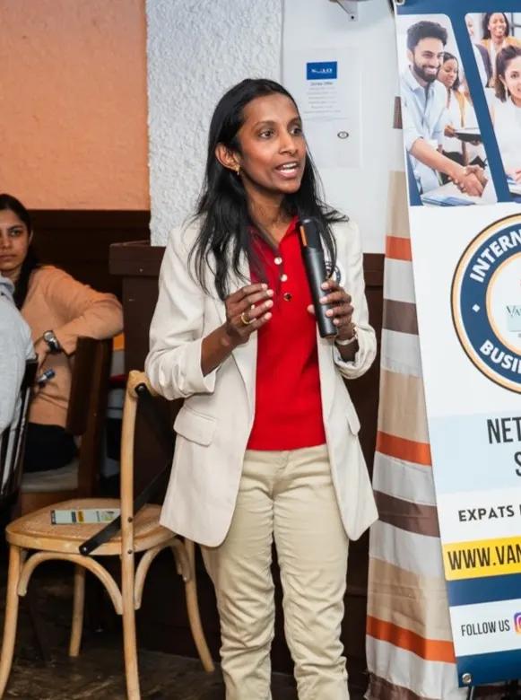 A woman in a cream blazer holds a microphone and speaks, standing next to a roll-up banner for an international business expo networking session.