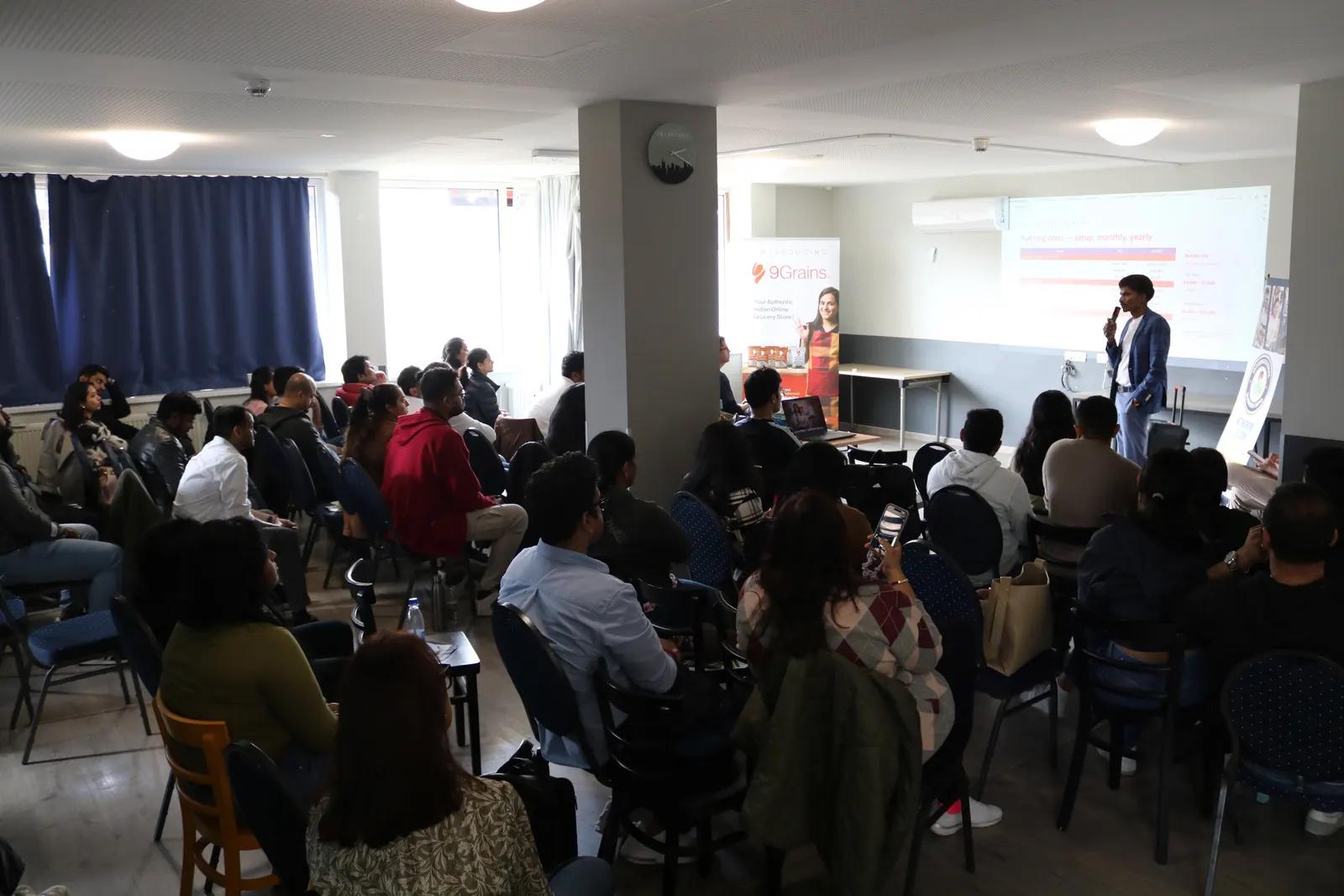 A speaker presents to a full room of seated attendees in a bright seminar room, with a slide screen and roll-up banner at the front.